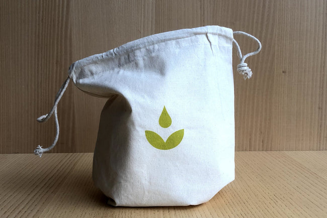 A natural cotton drawstring bag with a minimalist green leaf and water droplet logo, placed on a light wood surface against a matching wood panel background.
