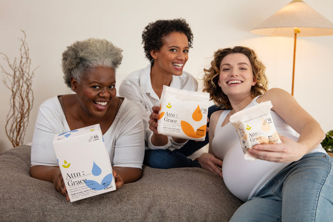 three women sitting on a bed holding up Attn: Grace bladder leak pads and liners